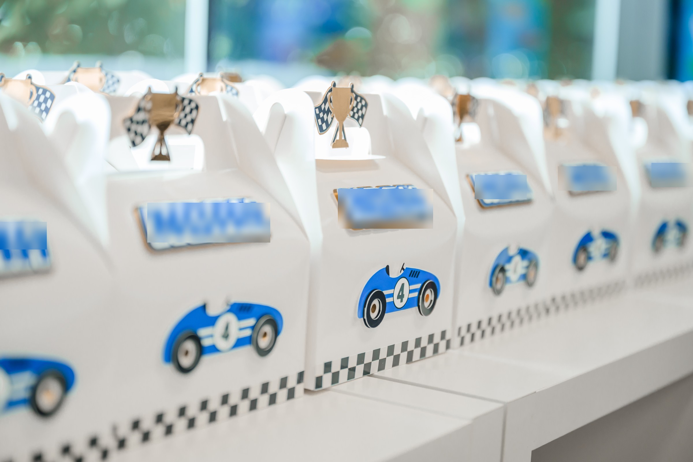Row of white race car birthday party favor boxes with blue vintage car number 4 graphics, trophy toppers, and personalized cookies