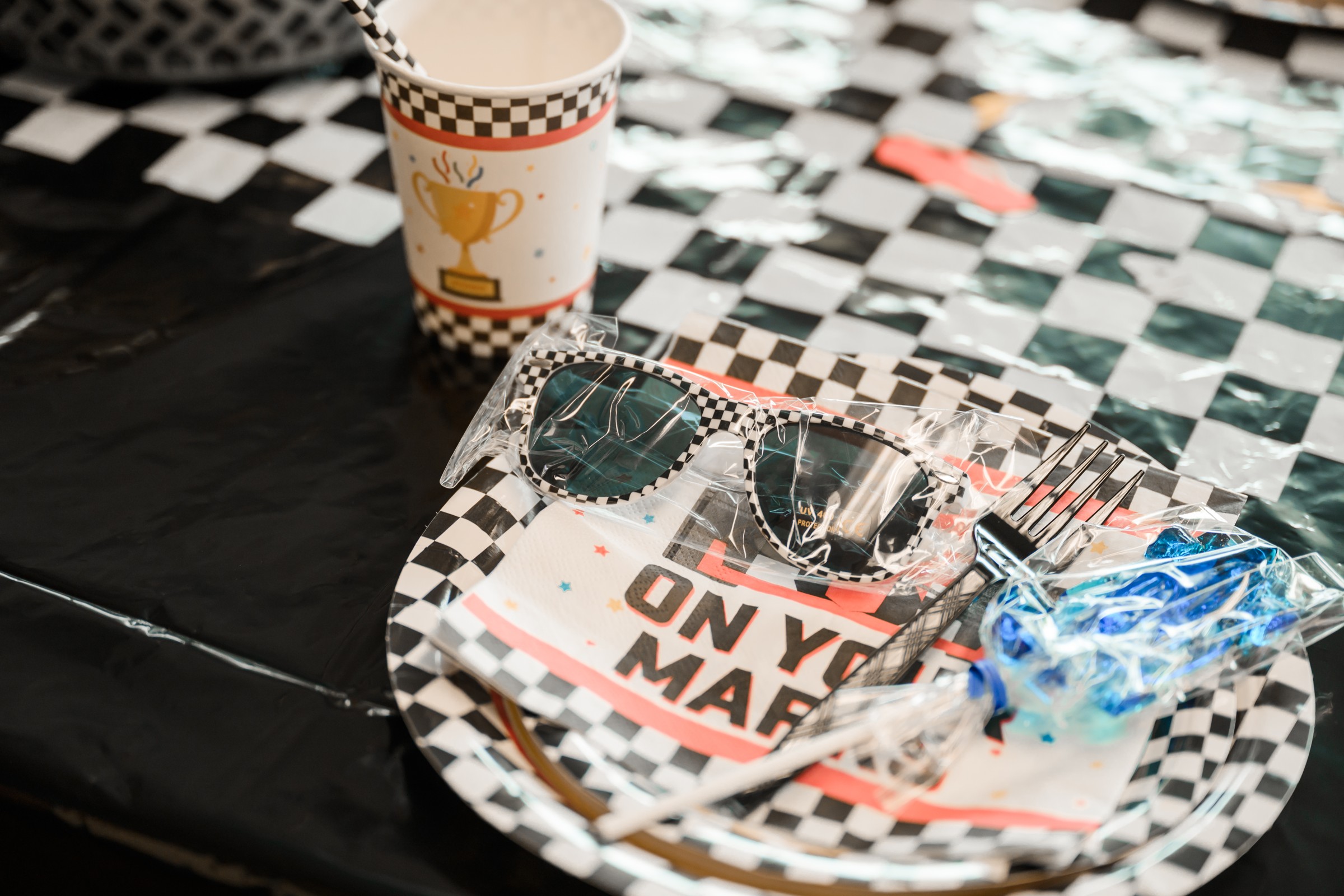 Race car birthday party place setting with checkered plate, On Your Mark napkin, checkered sunglasses favor, and trophy cup