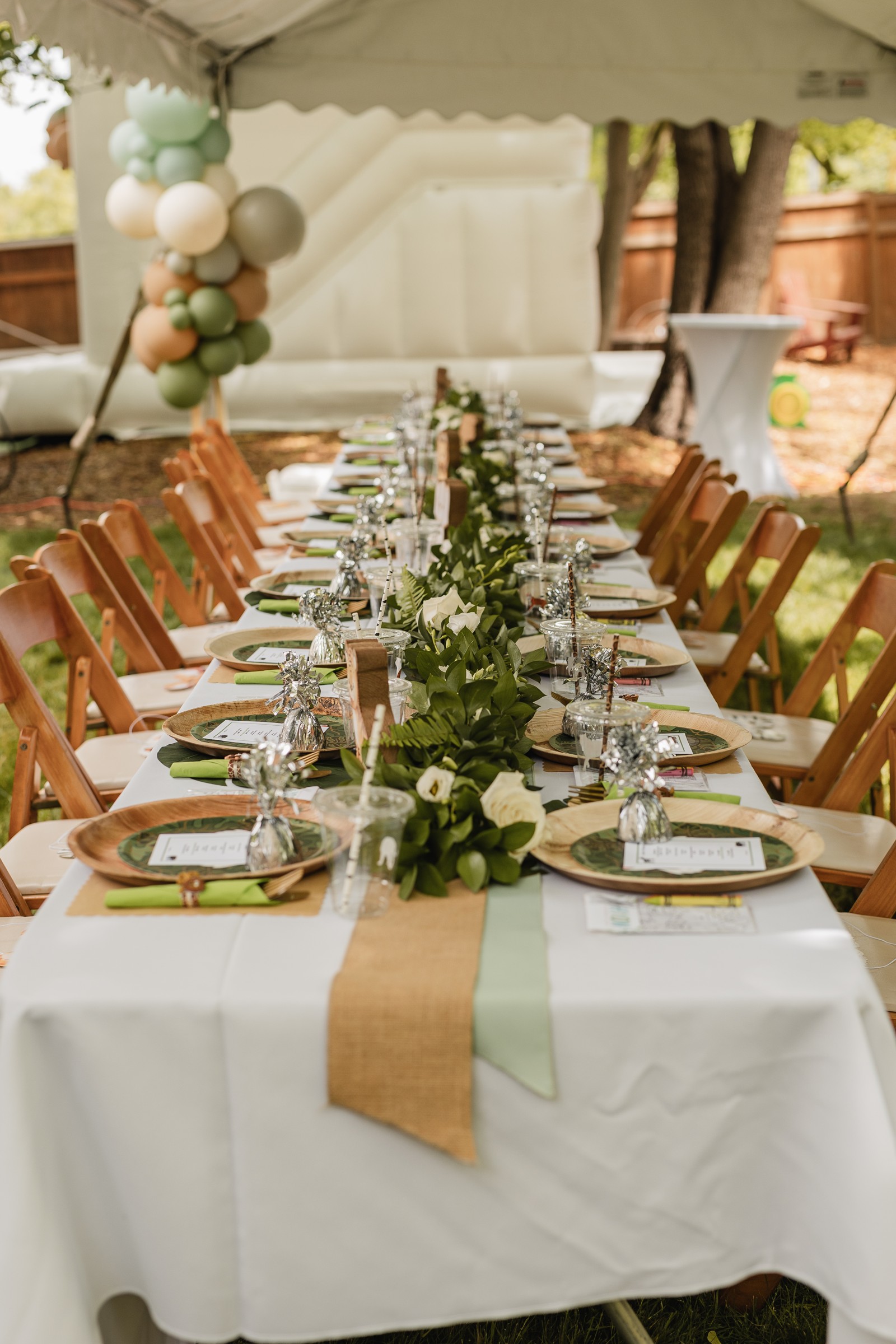 Outdoor safari birthday party banquet table set under a white tent with fern garland centerpiece and wooden charger plates